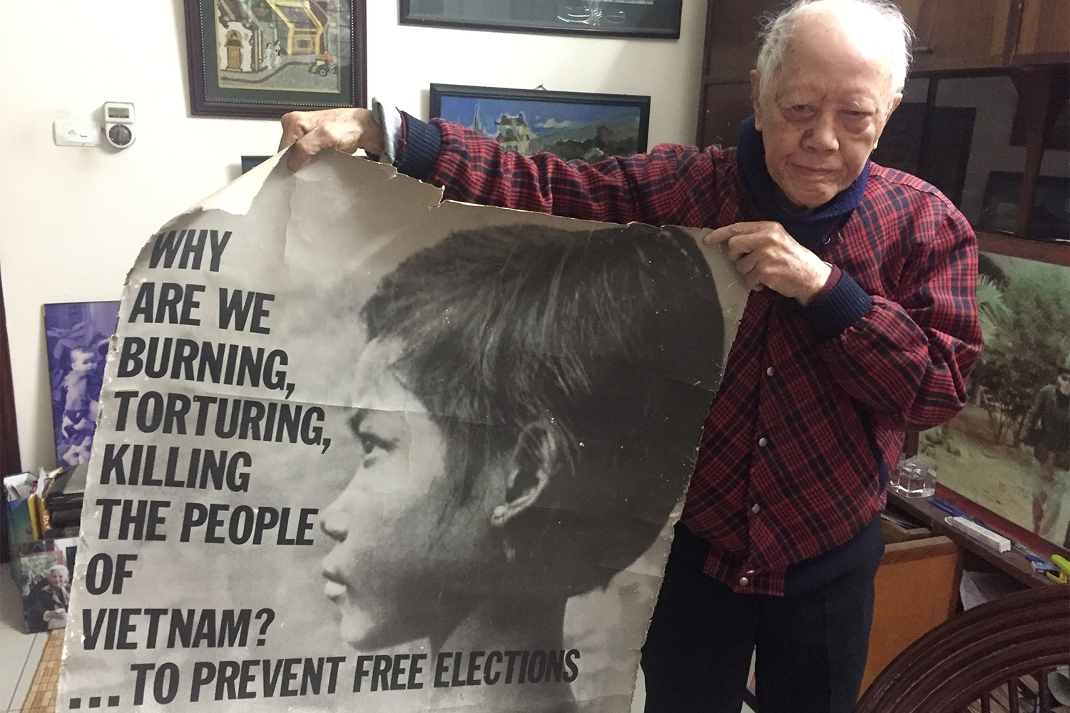 Hữu Ngọc chez lui, dans son bureau, à Hanoi. Il tient dans ses mains une affiche américaine d’époque, en faveur de la fin de la guerre au Vietnam.