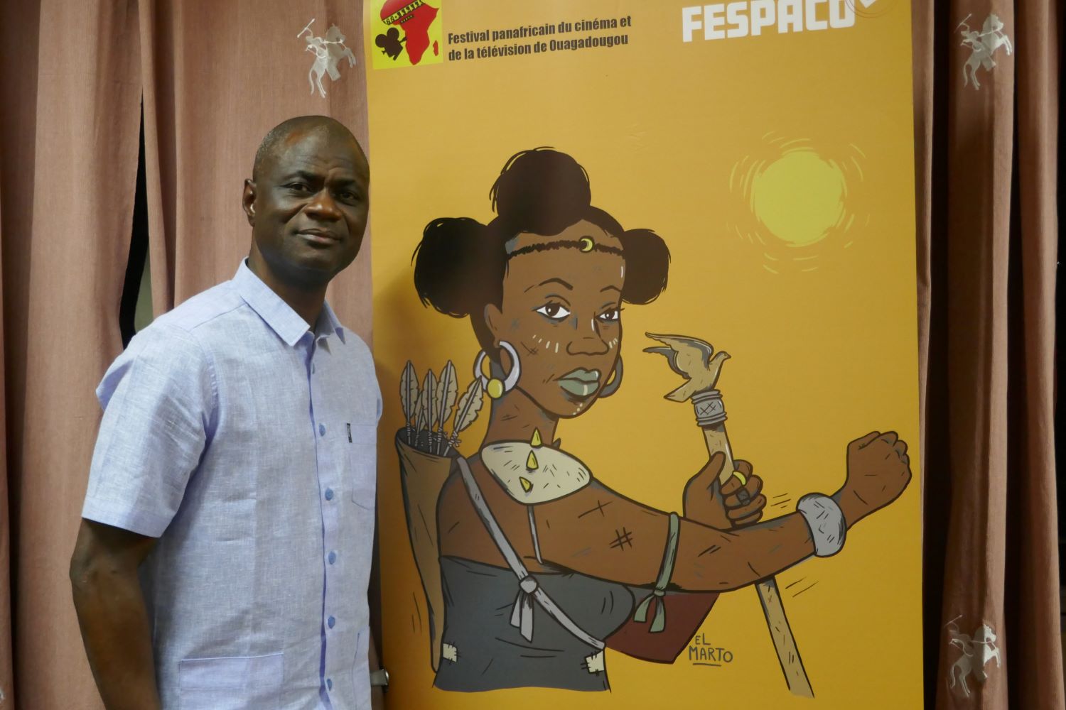 Alex Moussa SAWADOGO, à côté de l’affiche de cette édition du FESPACO, lance un message aux globe-reporters : « il faut étudier et aimer le cinéma pour voyager dans d’autres pays et les aimer » © Globe Reporters