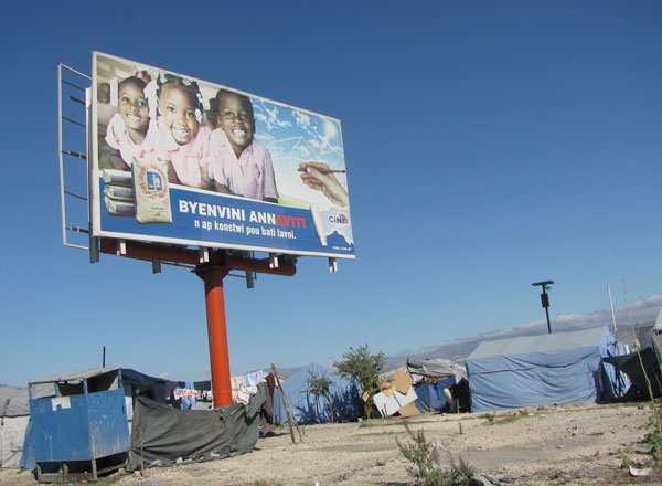 Nous passons à la hauteur de l’aéroport Toussaint Louverture. Une affiche souhaite « Bienvenue en Haïti ». Sous l’affiche, des toiles de tente. C’est un campement de gens dont le logement a été détruit par le tremblement de terre du 12 janvier 2010. Depuis cette date des dizaines de milliers de personnes vivent dans des conditions précaires à Port au Prince.